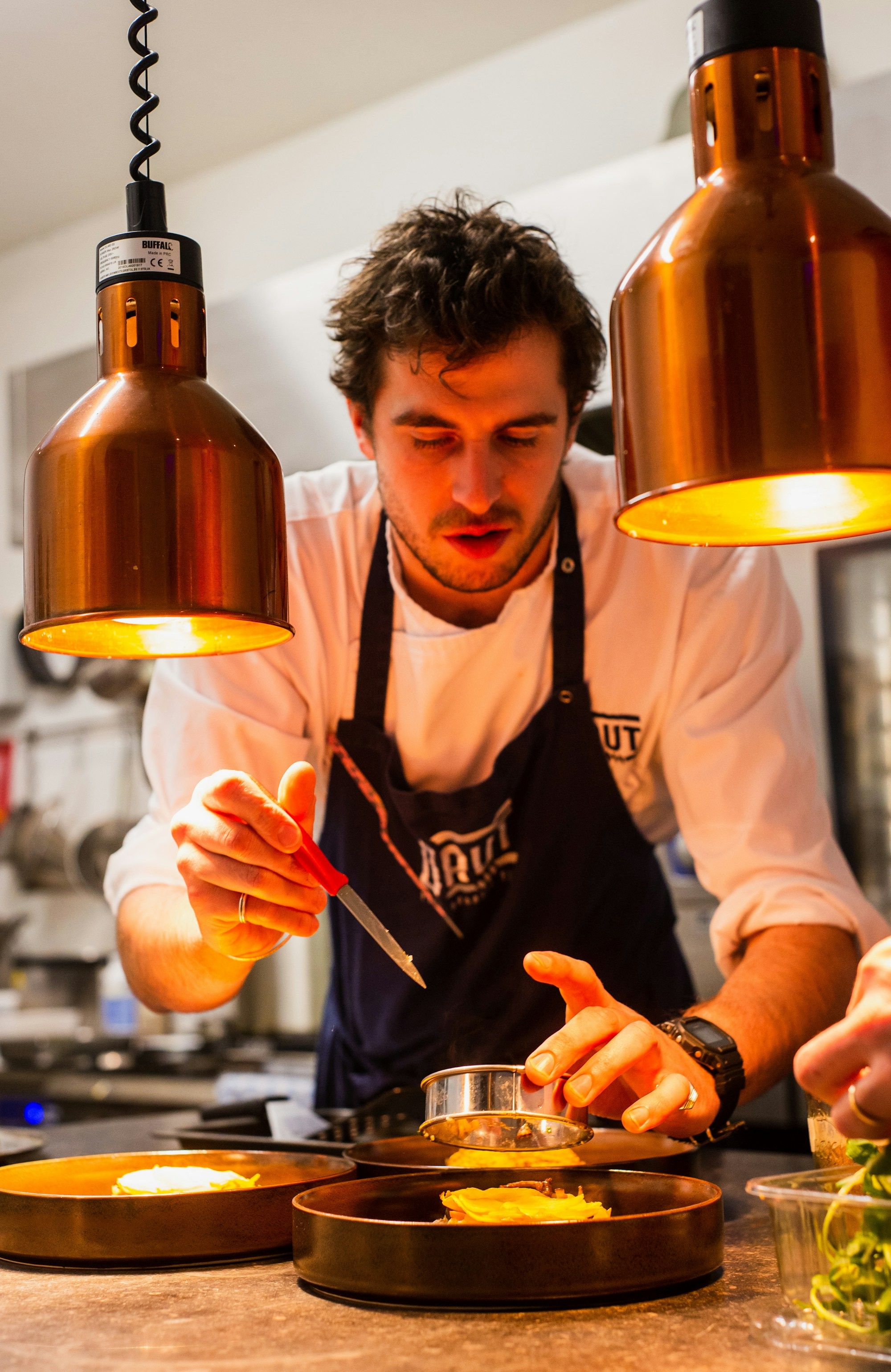Chef en tablier sous des lampes cuivrées, en train de dresser une assiette